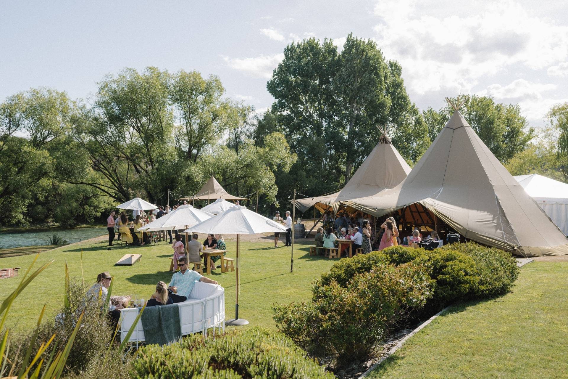 People are enjoying a sunny outdoor event with tents, tables, and umbrellas on a grassy area near a river