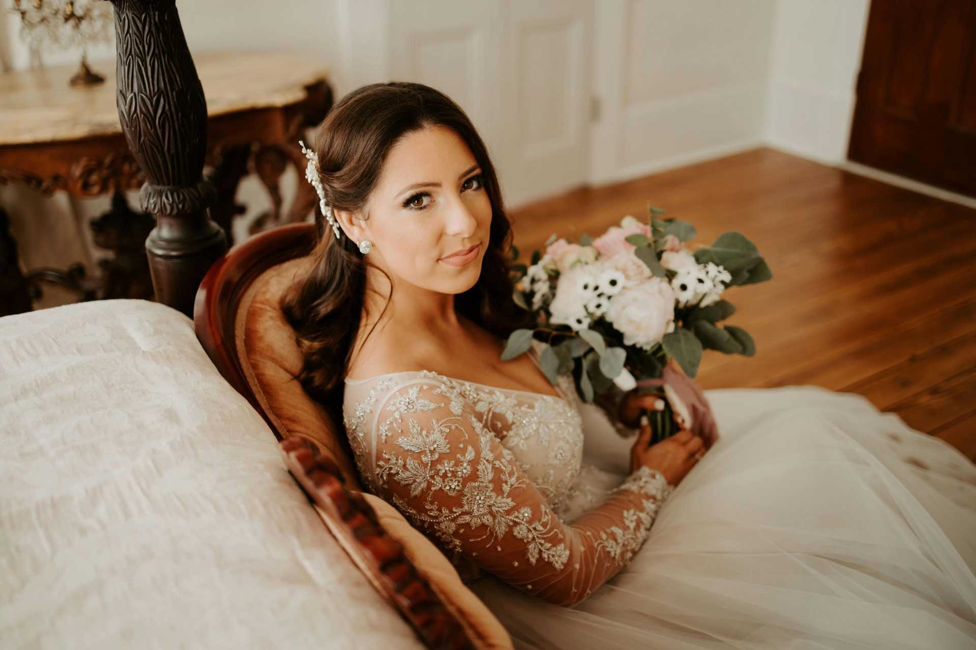 Bride in an elegant lace wedding dress sitting on a vintage chair holding a bouquet