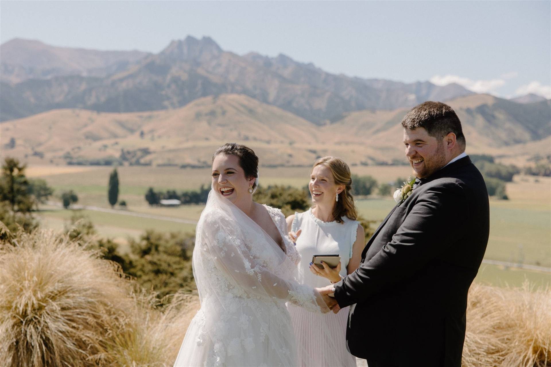 A bride and groom smiling and holding hands outdoors with an officiant and mountains in the background